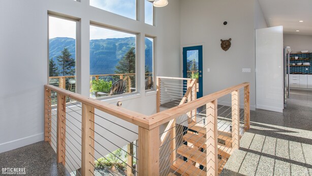 A modern interior with large windows offering mountain views, a wooden staircase with a glass railing, and a kitchen area. - Dream Views & Chef's Kitchen-Modern, Mountain Home w/ Hot Tub (Anchorage)