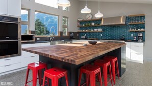 A modern kitchen with a large island, red bar stools, white cabinets, and a view of mountains through large windows. - Dream Views & Chef's Kitchen-Modern, Mountain Home w/ Hot Tub (Anchorage)