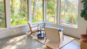 A room with large windows offering a view of a forested area. There are two chairs and a small table. - Dream Views & Chef's Kitchen-Modern, Mountain Home w/ Hot Tub (Anchorage)