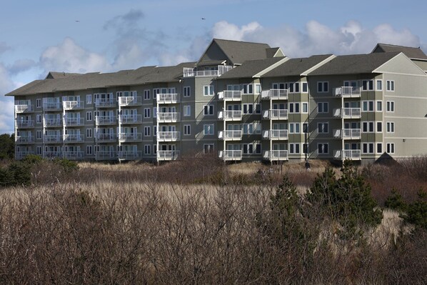 A multi-story residential building with balconies, a flat roof, and a sandy foreground.