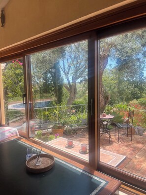 Ein Zimmer mit großem Fenster mit Blick auf einen Garten, inklusive Tisch, Stühlen, einem Korbwägelchen und einem kleinen Tisch mit Kerze.