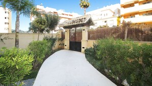 A pathway leading to a gated entrance with a traditional archway, surrounded by greenery and palm trees, with buildings in the background. - 1081 Apartment Poseidon (Nerja)