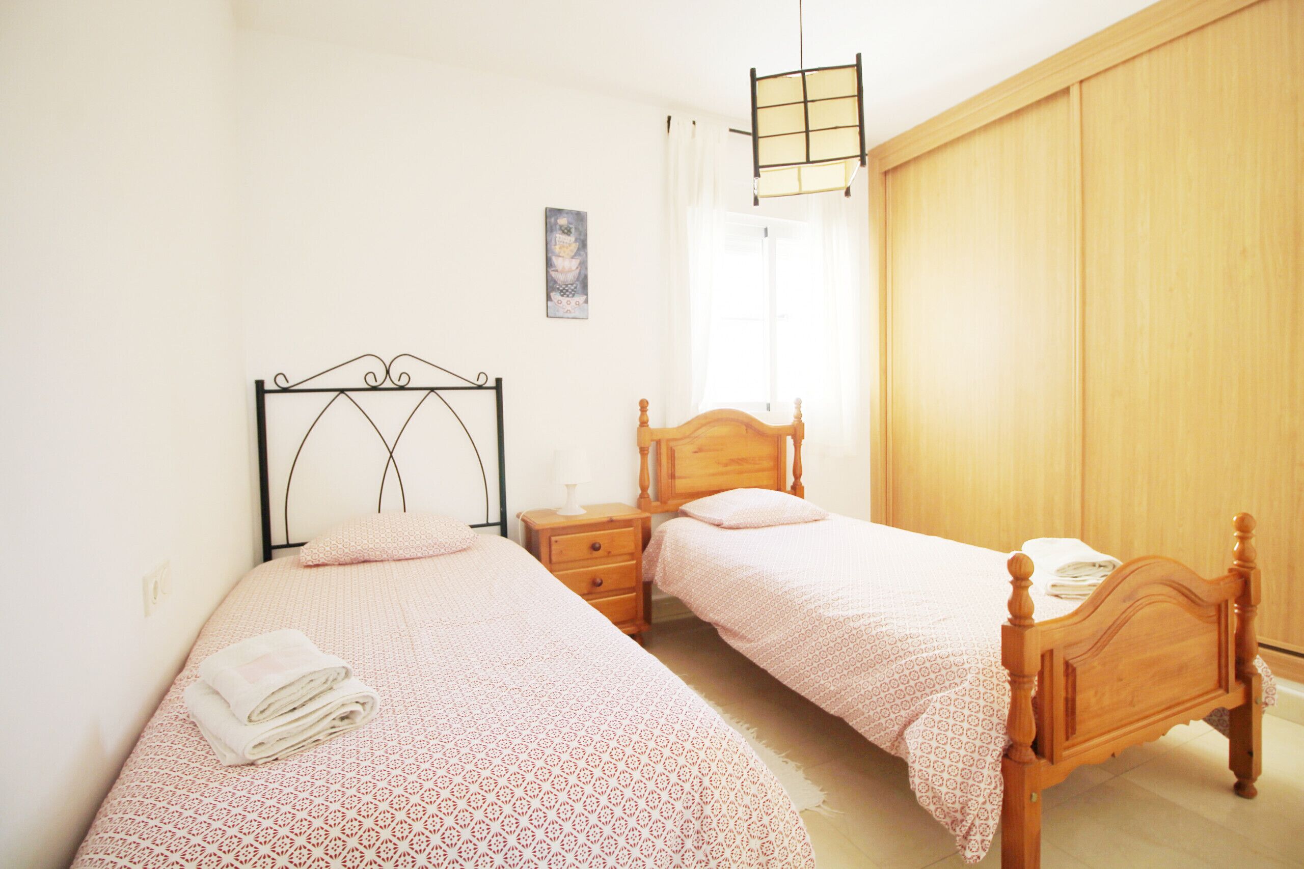 A bedroom with two single beds, a bedside table with a lamp, a hanging light fixture, and a wooden wardrobe.