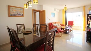 A dining area with a glass table and wooden chairs, a living room with a sofa and armchairs, and a window with curtains. - 1081 Apartment Poseidon (Nerja)