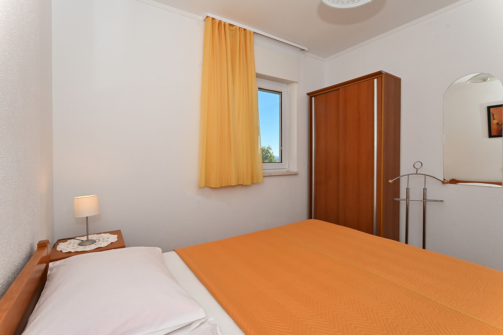 Habitació d'hotel amb un llit, una finestra amb cortines grogues, un armari de fusta, una tauleta de nit amb una làmpada i un mirall a la paret.