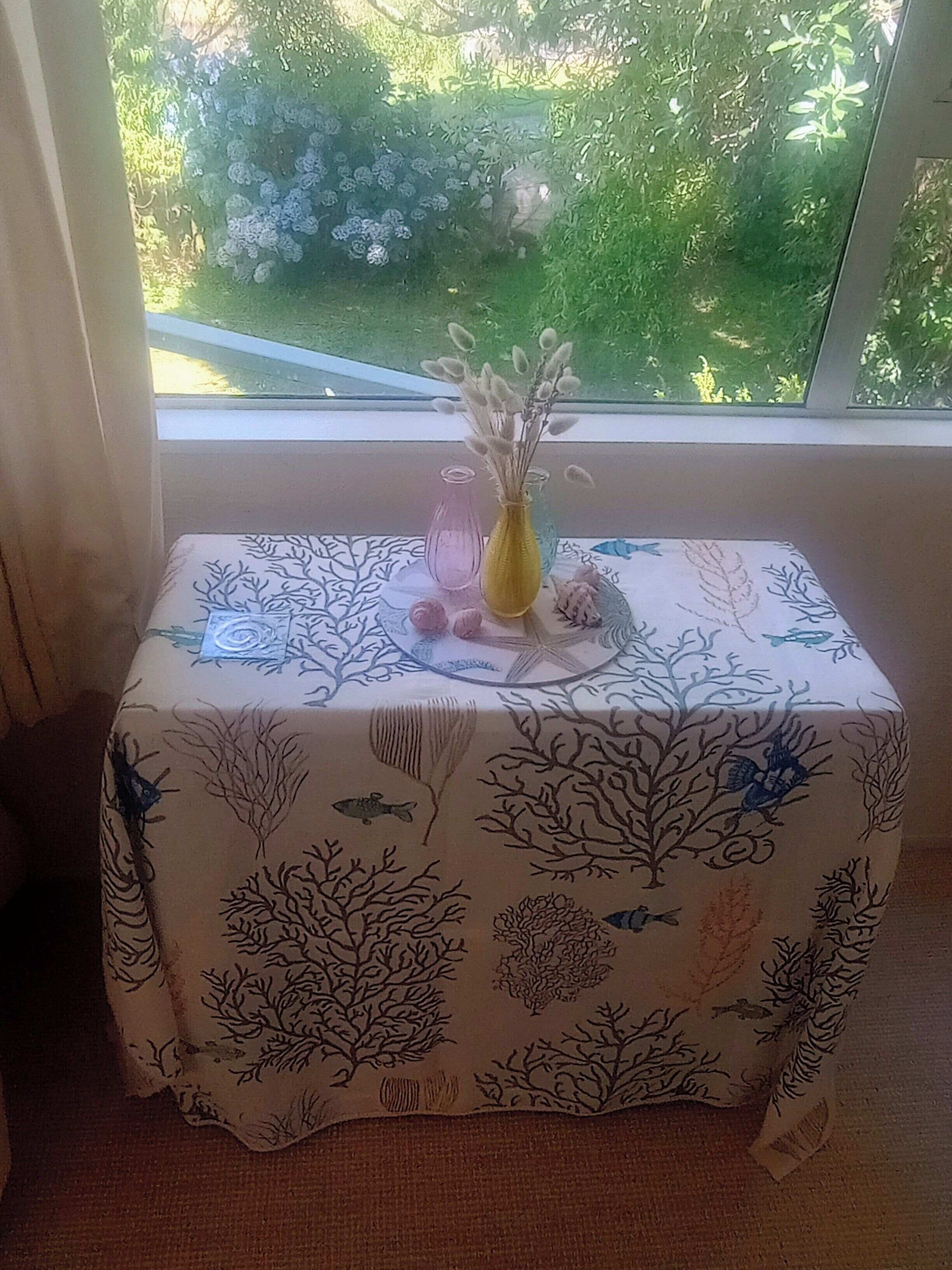 Un tavolino con tovaglia floreale, due vasi e un oggetto decorativo su un davanzale.