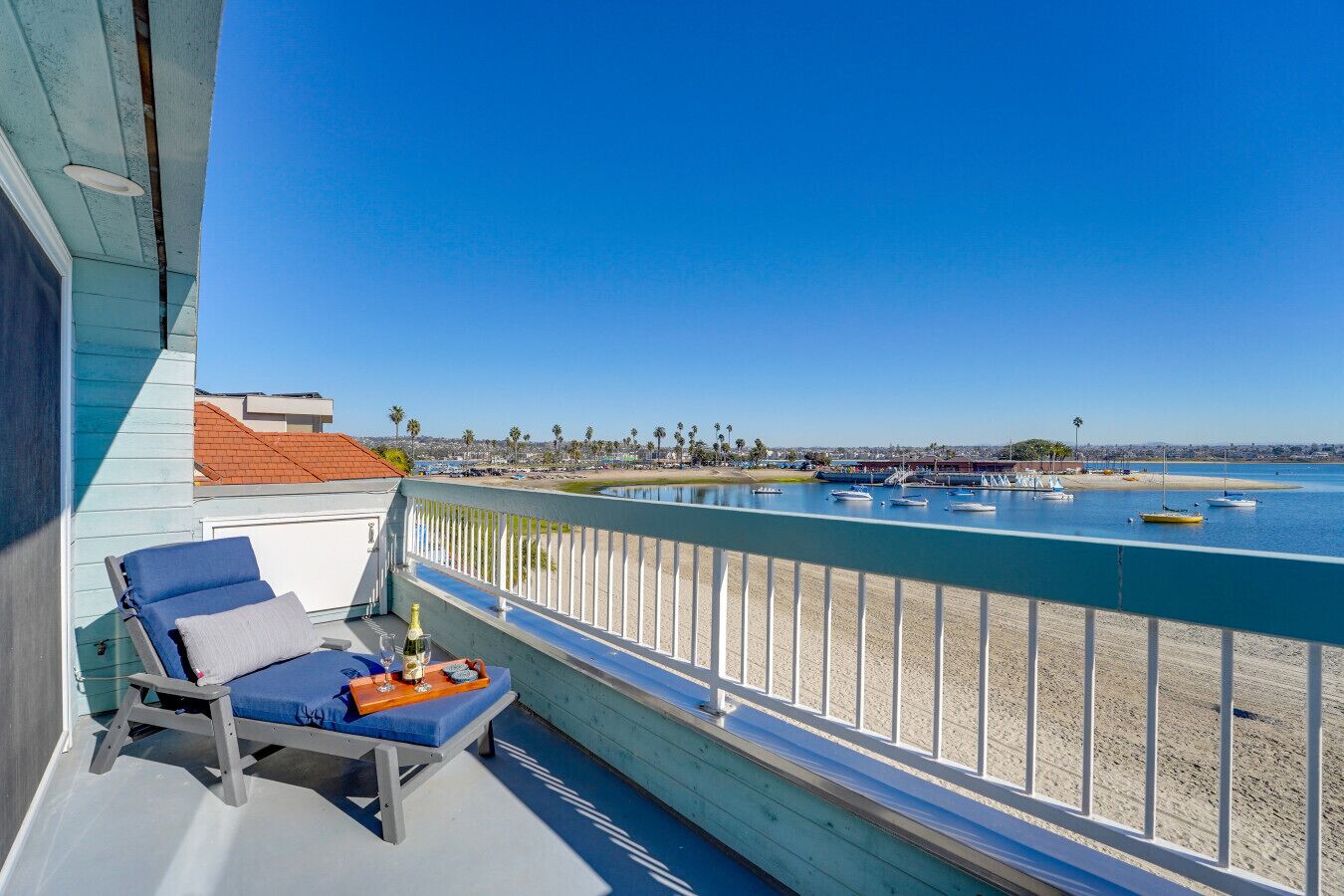 A balcony with a blue cushioned chair, a small table with a bottle and glasses, and a view of the beach and boats.
