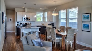 A modern kitchen with white cabinets, stainless steel appliances, and a dining area with wicker chairs and a wooden table. - Sea Turtle Cottage - Oyhut Bay Seaside Village (Ocean Shores)