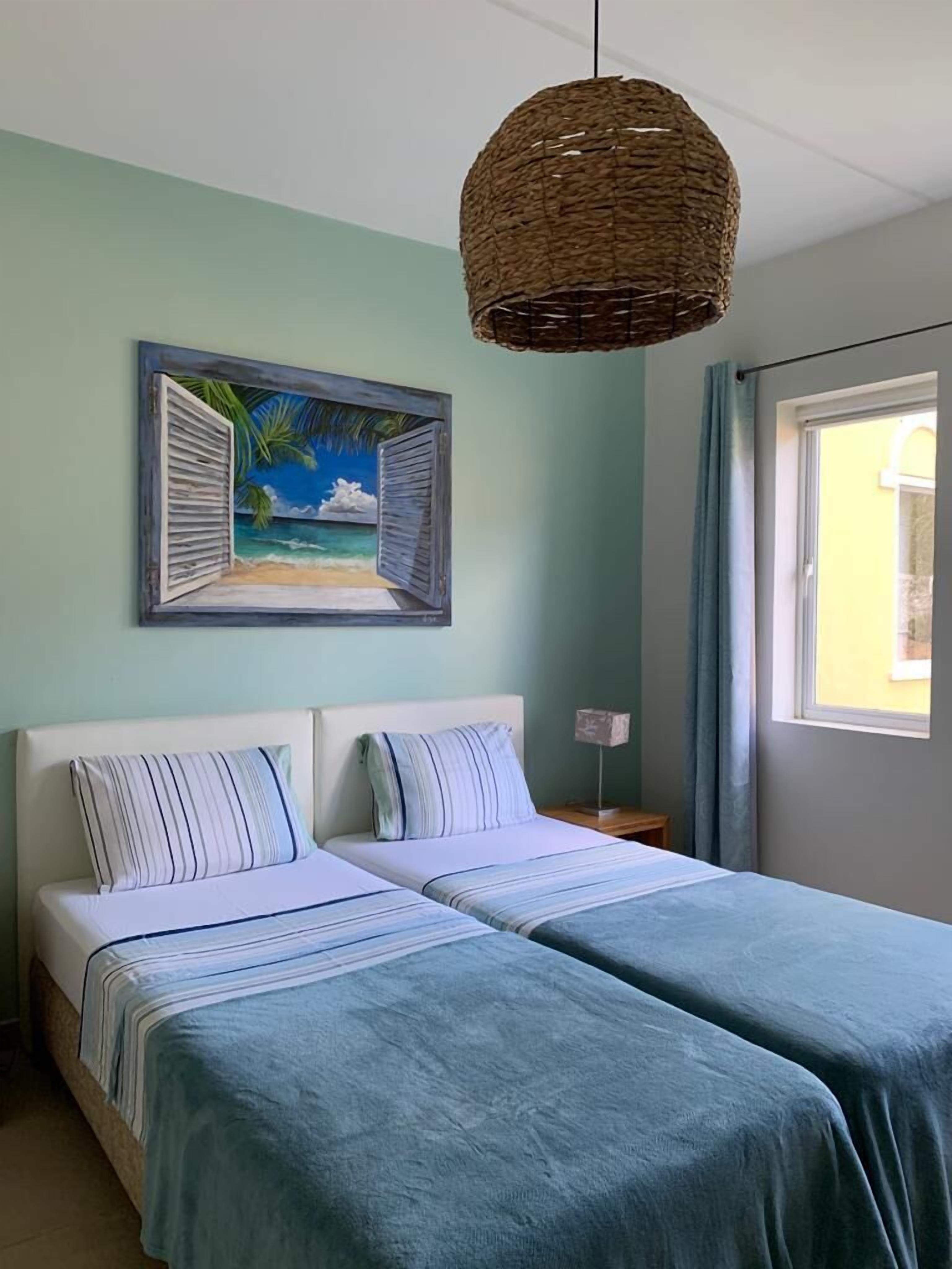 Un dormitorio con dos camas, un cuadro de una escena de playa, una lámpara colgante de mimbre y una ventana con cortinas.