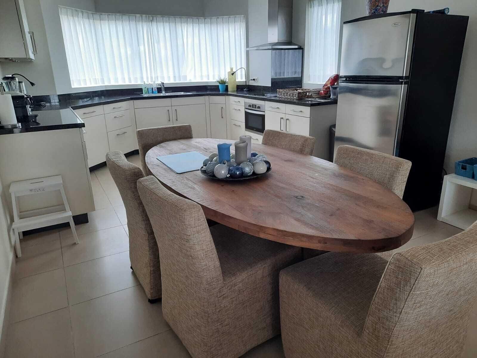A modern kitchen with a wooden dining table, beige upholstered chairs, and a stainless steel refrigerator.