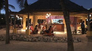 Un bar en la playa con terraza, techo de paja y palmeras.