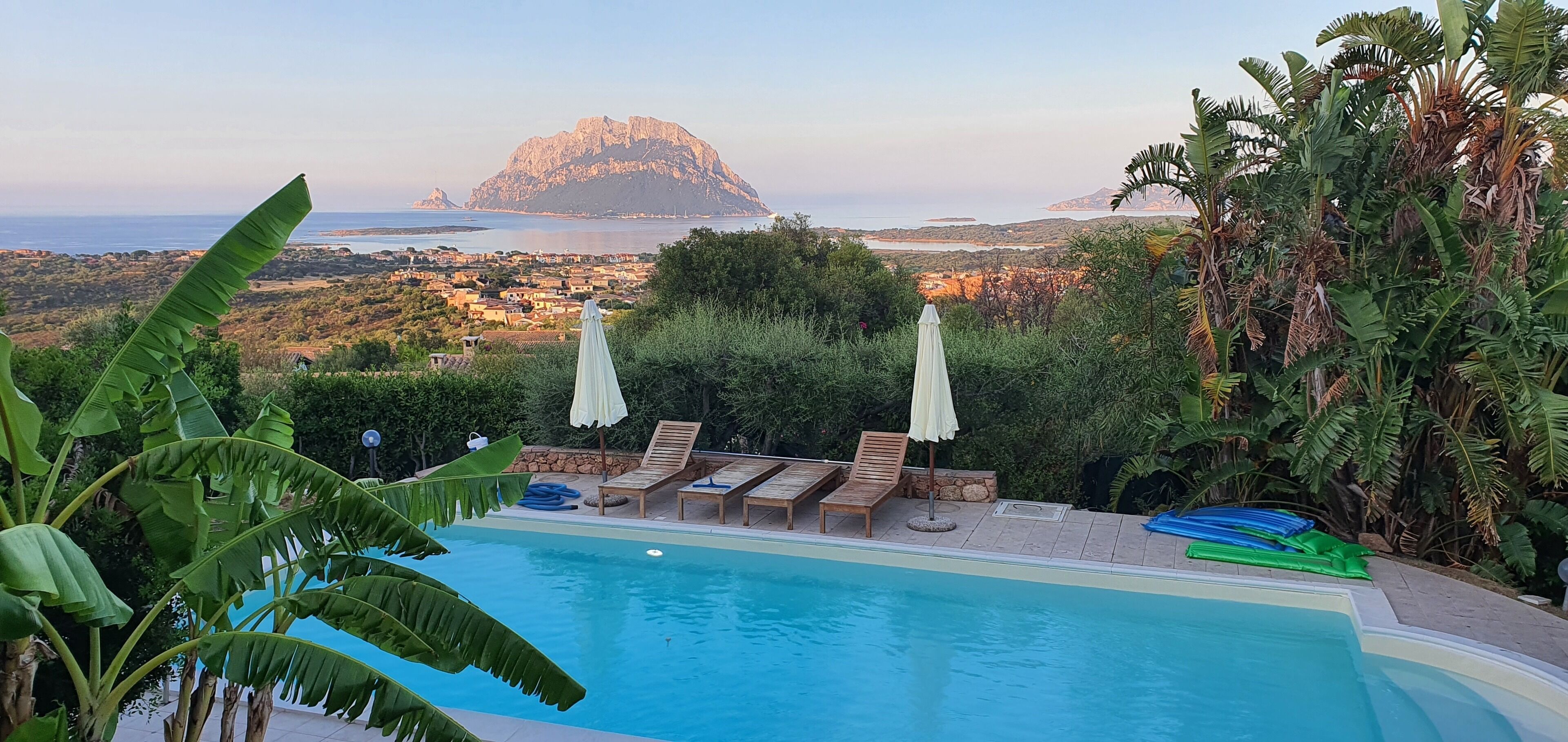 Un área de piscina con sillones de descanso y sombrillas, rodeada de exuberante vegetación y con vistas a un paisaje pintoresco que incluye una montaña imponente y la costa.