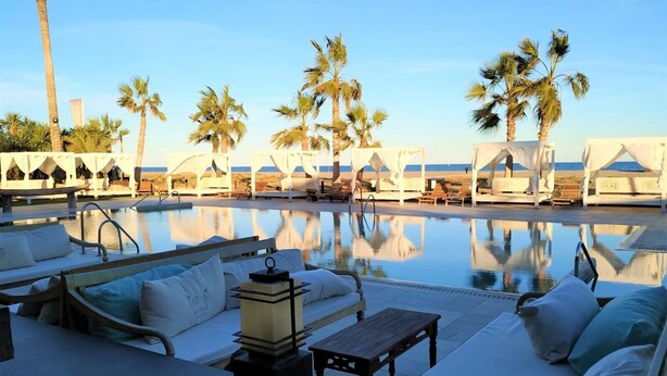 Un espace aménagé au bord de la piscine, avec des chaises longues et des parasols, une piscine dans laquelle se reflètent les palmiers, et une vue sur l’océan.