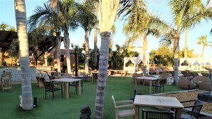 Un espace extérieur aménagé avec des tables et des chaises, entouré de palmiers et bordé d’une piscine.