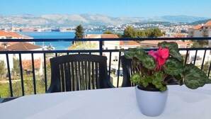 Uma varanda com mesa, cadeira e um vaso com flores cor-de-rosa. É visível uma paisagem com uma vila costeira e montanhas ao fundo.