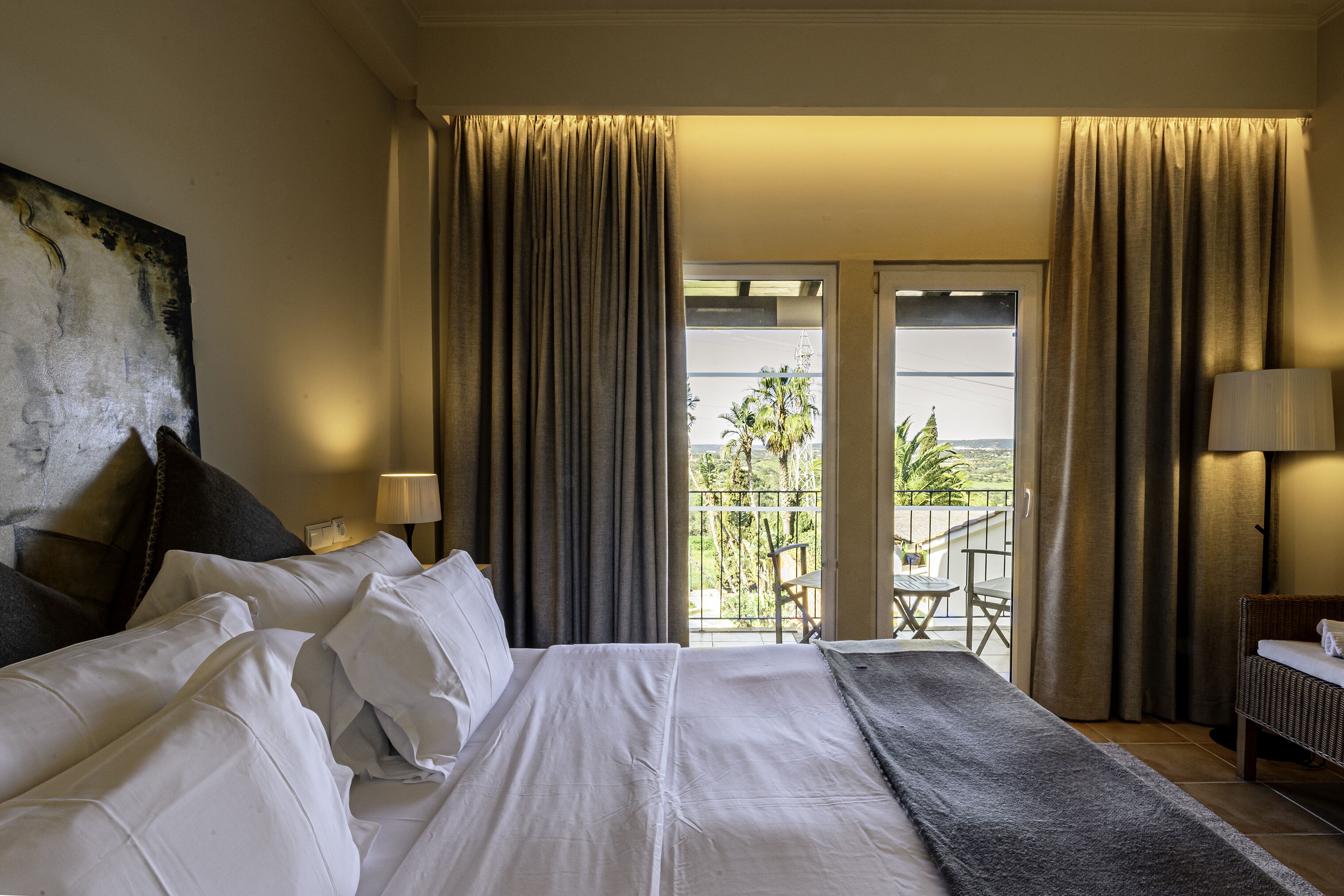 Habitación de hotel con una cama grande, un balcón con mesa y sillas, y cortinas.
