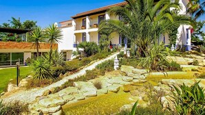 Un edificio de varios pisos con balcones, rodeado de plantas tropicales y un camino de piedra que conduce a una fuente de agua.