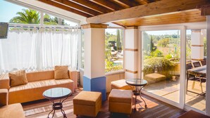 Un patio iluminado por el sol con techo de madera, un sofá color beige, dos otomanas y una mesa de vidrio.