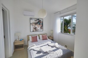 Une chambre à coucher comprenant un lit, une table de chevet, un climatiseur fixé au mur, une fenêtre avec des rideaux et un luminaire au plafond.