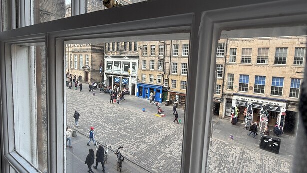 A view from a window overlooking a cobblestone square with historic buildings, a shop named "Crest of Edinburgh," and people walking.