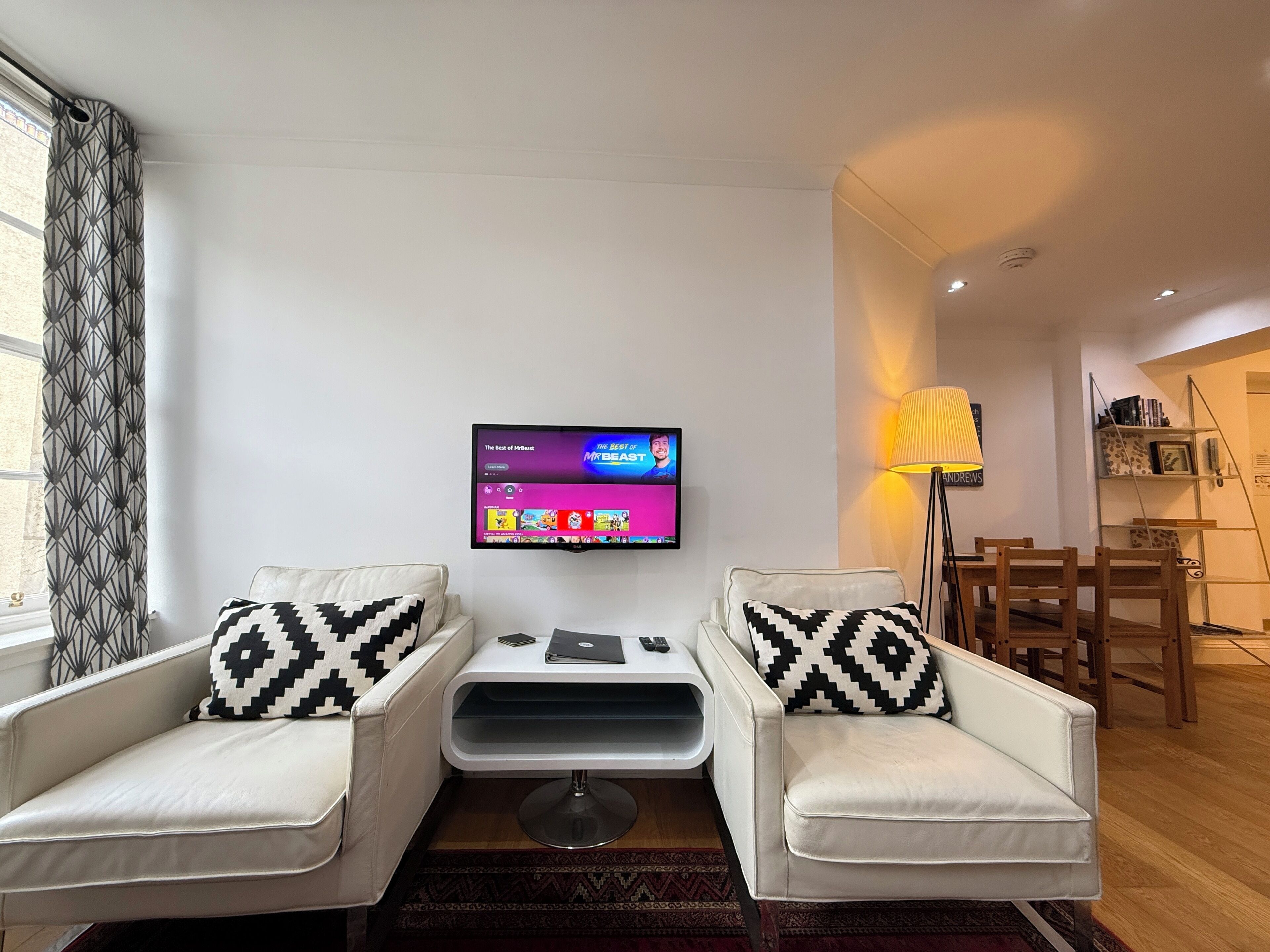 Een moderne woonkamer met twee witte fauteuils, een flatscreen televisie, een klein tafeltje en een lamp.