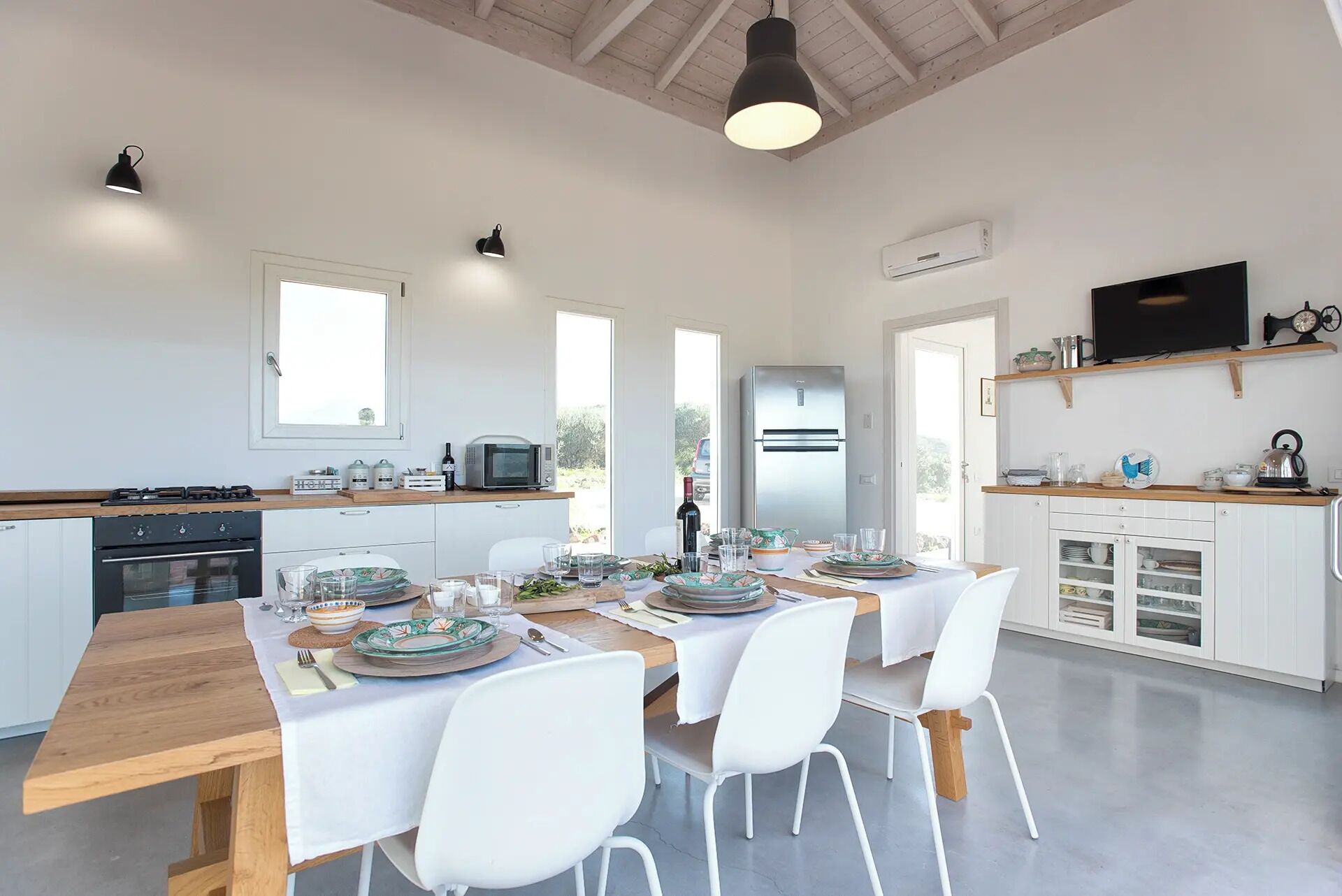 A modern kitchen with a dining area, white cabinetry, a built-in oven, a microwave, a refrigerator, and a television on a shelf.