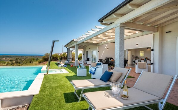 Zona junto a la piscina con sillones de descanso, una pérgola y vista al océano.