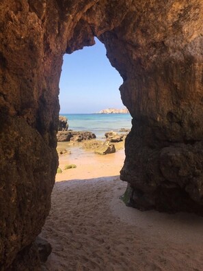 Um arco natural de rocha que dá para uma praia arenosa, com águas azuis cristalinas e formações rochosas ao longe.