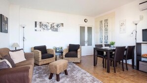 A living room with a sofa, two wicker chairs, a coffee table, and a dining table with chairs. There is a clock on the wall and a TV on the right. - Ocean view - 150 m beach - refurbished 4 to 6 places in Praia da Rocha 95 M2 (Portimão)