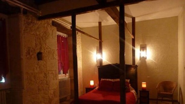 Un llit amb cortines i pilars, un tamboret i dues làmpades de nit en una habitació amb parets de pedra i una finestra amb cortines vermelles.