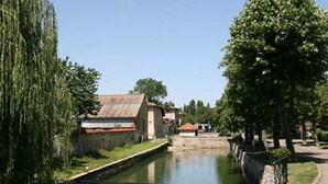 Un canal amb un pont de pedra, un edifici petit amb teulada vermella i arbres alineats al camí.