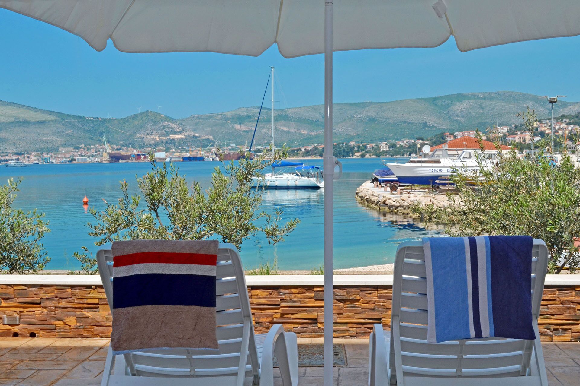 Vista para o mar com duas espreguiçadeiras sob um grande guarda-sol, com vista para um porto cheio de barcos e uma cidade ao longe.
