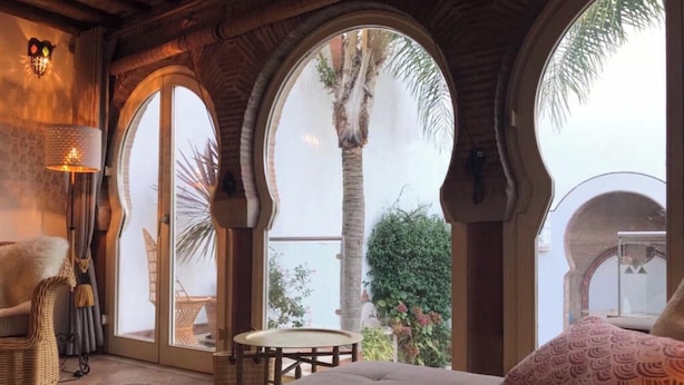 Une chambre avec un grand lit, une chaise, une table et une vue sur un palmier, visible à travers des fenêtres en arc de cercle.