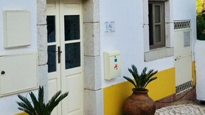 Un bâtiment avec un toit recouvert de tuiles, une façade blanche et une partie inférieure jaune. Deux plantes en pot sont placées devant l’entrée.