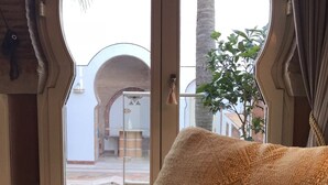 Une pièce dotée d’une grande fenêtre en arc de cercle, d’un canapé agrémenté de coussins à motifs et offrant une vue sur un espace extérieur avec des palmiers et un bâtiment aux arches.