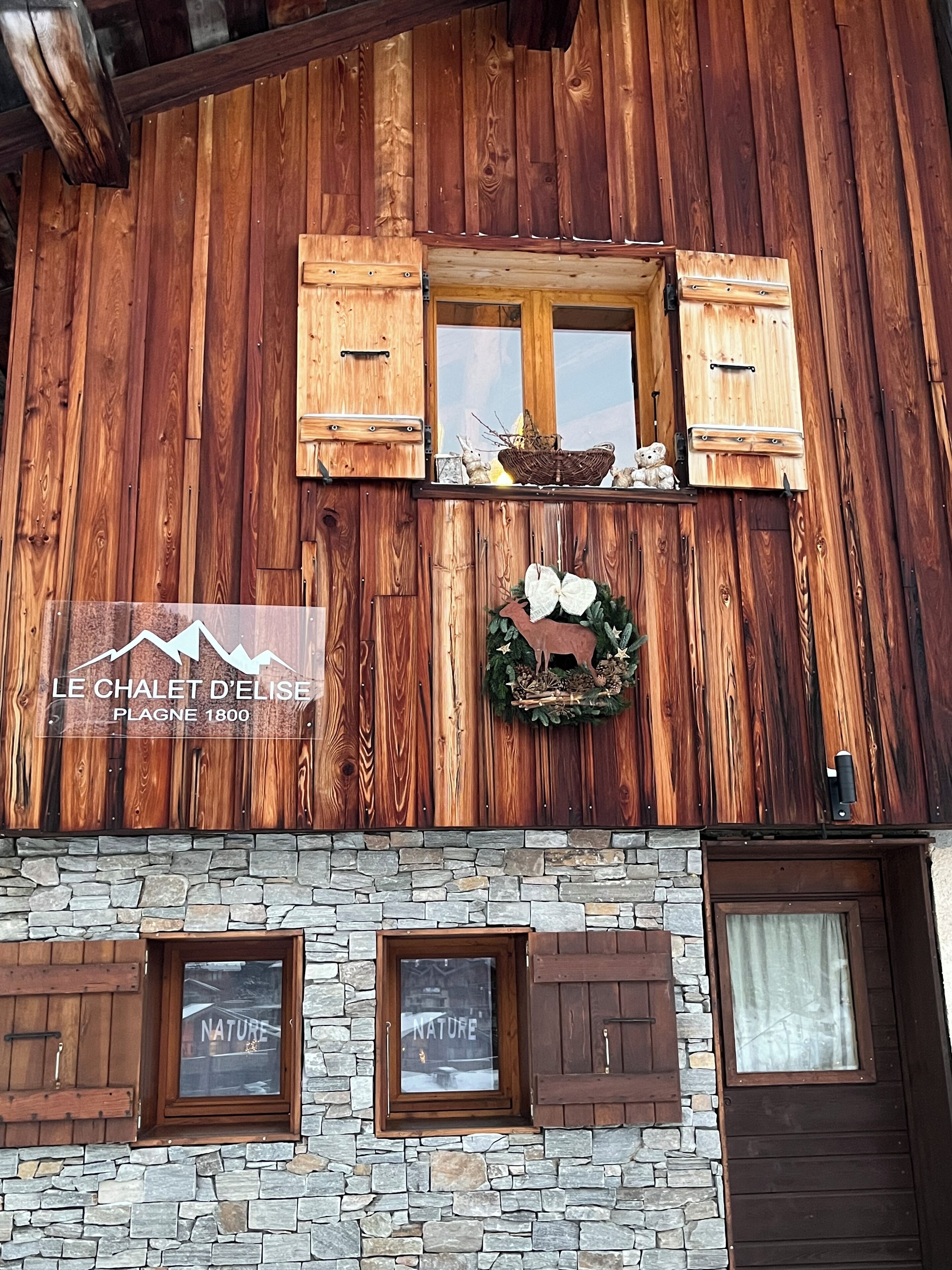 Ein Holzgebäude mit einer Steinaußenwand, Holzläden und einem Schild mit der Aufschrift 'LE CHALET D'ELISE PLAGNE 1800'.