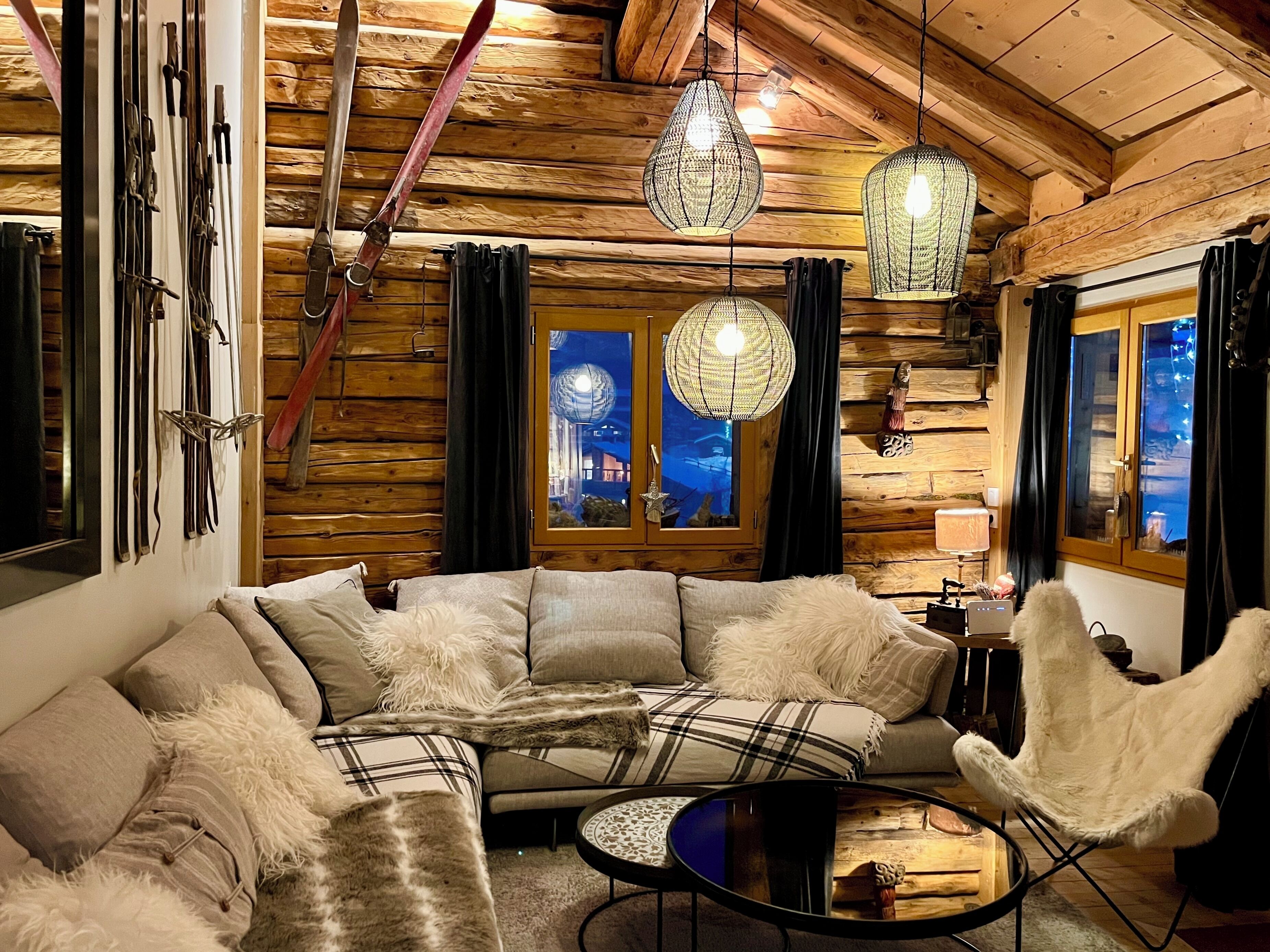 A cozy cabin interior with a large L-shaped sofa, a round coffee table, and a white armchair. There are skis and ski poles on the wall, and a large window with a view of a building at night.