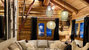 A cozy cabin interior with a large L-shaped sofa, a round coffee table, and a white armchair. There are skis and ski poles on the wall, and a large window with a view of a building at night. - Chalet Plagne (1800) (Mâcot-la-Plagne)