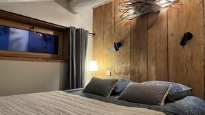 A bedroom with a large bed, a window, wooden walls, and a wall-mounted light fixture. - Chalet Plagne (1800) (Mâcot-la-Plagne)