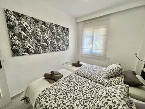 Une chambre avec deux lits, une grande œuvre d’art à motifs floraux accrochée au mur, une fenêtre avec des stores et une table de chevet avec une lampe.