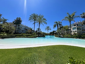 Une piscine à l’eau d’un bleu cristallin, entourée de pelouses bien entretenues et de palmiers, avec des immeubles à plusieurs étages en arrière-plan.