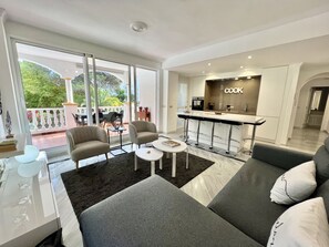 A modern living room with a sofa, a coffee table, and a dining area with chairs. There is a kitchen area with a 'COOK' sign visible.