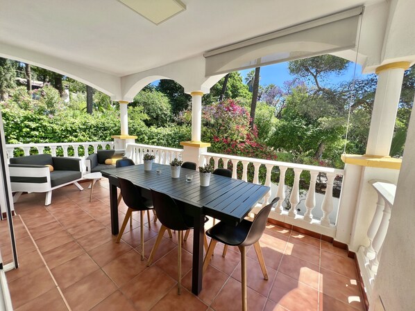 Une terrasse couverte, agrémentée d’une table et de chaises, entourée d’une balustrade blanche et d’une végétation luxuriante.