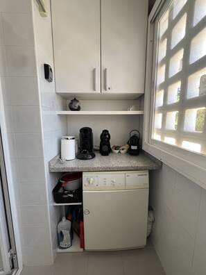 A compact kitchen with white cabinets, a countertop with coffee makers, a dishwasher, and a window with a grid pattern.
