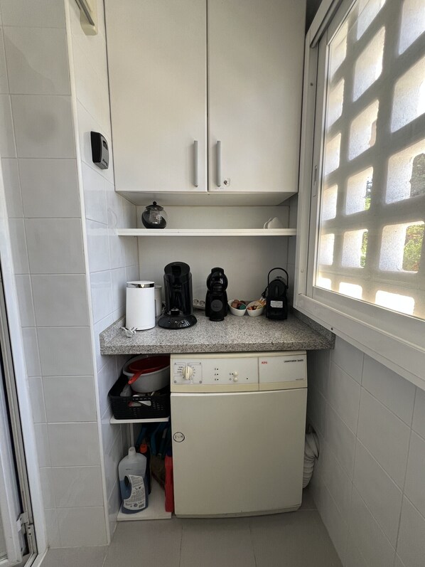 Eine kompakte Küche mit weißen Schränken, einer Arbeitsplatte mit Kaffeemaschinen, einem Geschirrspüler und einem Fenster mit Gittermuster.