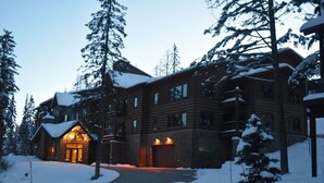 Un amplio edificio de estilo cabaña de troncos con numerosas ventanas y un techo cubierto de nieve, rodeado de altos pinos.