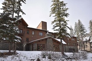 Une grande cabane en rondins de bois, dotée d’une cheminée en pierre, entourée d’arbres couverts de neige.