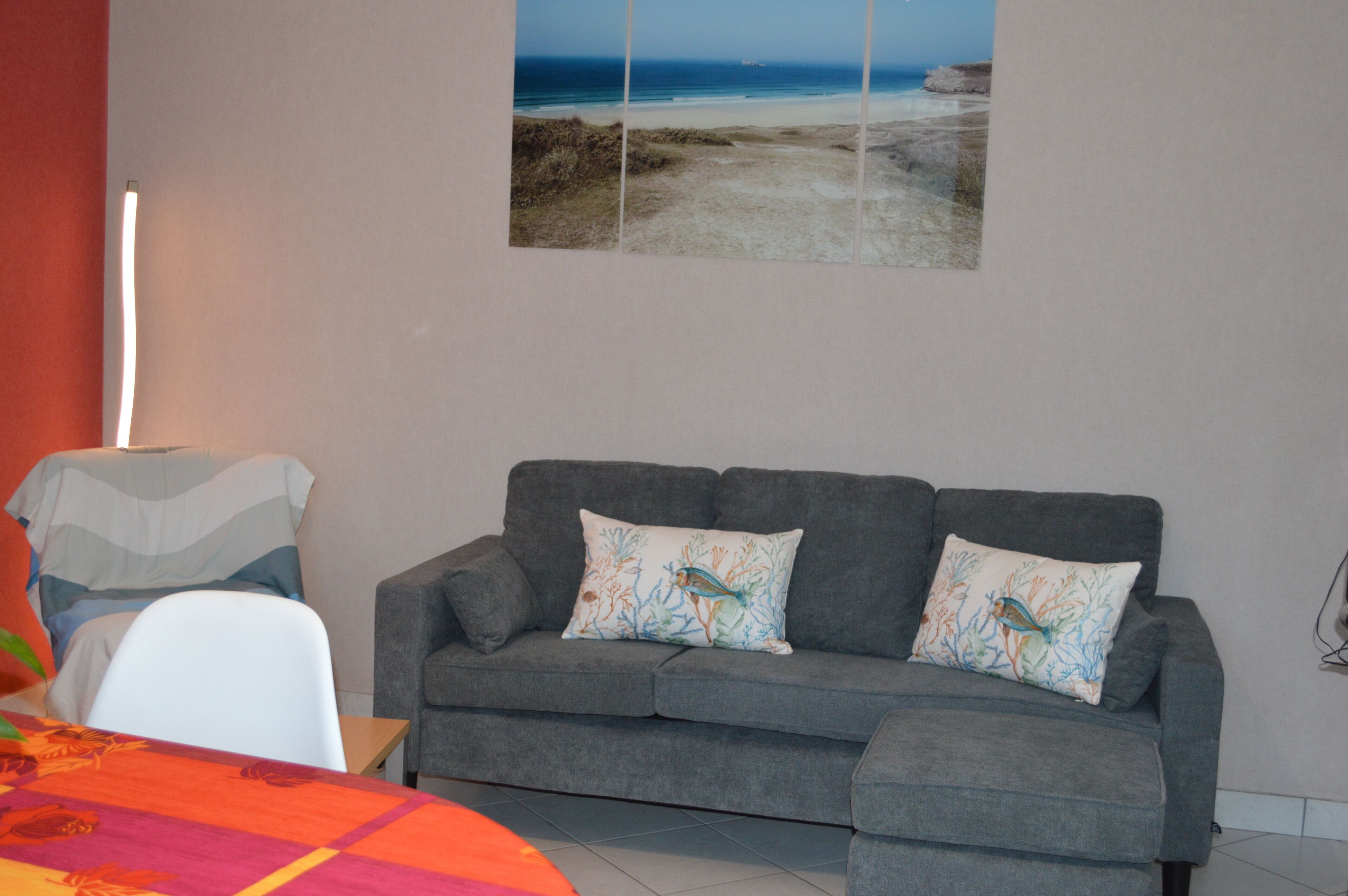 Un salon avec un canapé gris, deux coussins décoratifs, une table recouverte d’une nappe colorée et un tableau représentant une plage, accroché au mur.