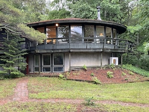 A modern, two-story wooden cabin with large windows and a balcony, surrounded by a forested area.