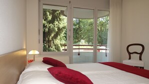 Um quarto com uma cama grande, uma janela com vista para árvores, uma cadeira e uma mesa de cabeceira com abajur.